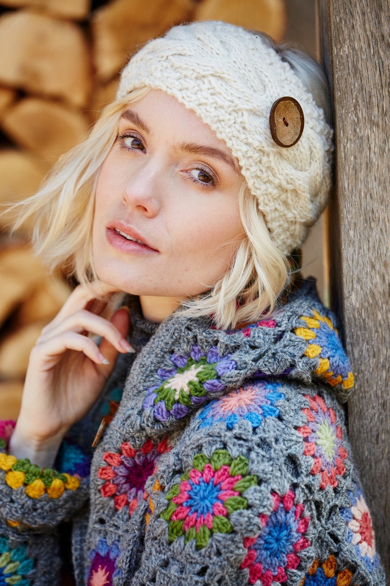 May include: A woman wearing a white crocheted headband with a wooden button and a grey crocheted sweater with colourful floral designs.