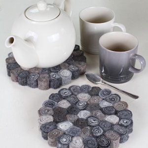 May include: Two grey and white felt coasters with a circular design. The coasters are made of rolled felt and are arranged in a circular pattern. A white teapot and two mugs are sitting on the coasters.