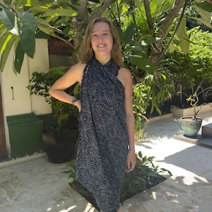 May include: A woman wearing a black and white patterned sarong dress. The dress has a halter neck style and is made of a lightweight fabric. The woman is standing in front of a lush green tropical garden.