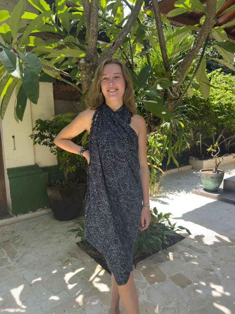 May include: A woman wearing a black and white patterned sarong dress. The dress has a halter neck style and is made of a lightweight fabric. The woman is standing in front of a lush green tropical garden.