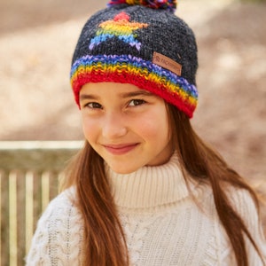 May include: A child wearing a gray knitted hat with a rainbow star design and a colorful pom-pom on top. The hat has a red, orange, yellow, green, blue, indigo, and violet stripe around the brim. The hat has a brown leather label with the text "Pachamama" on it.