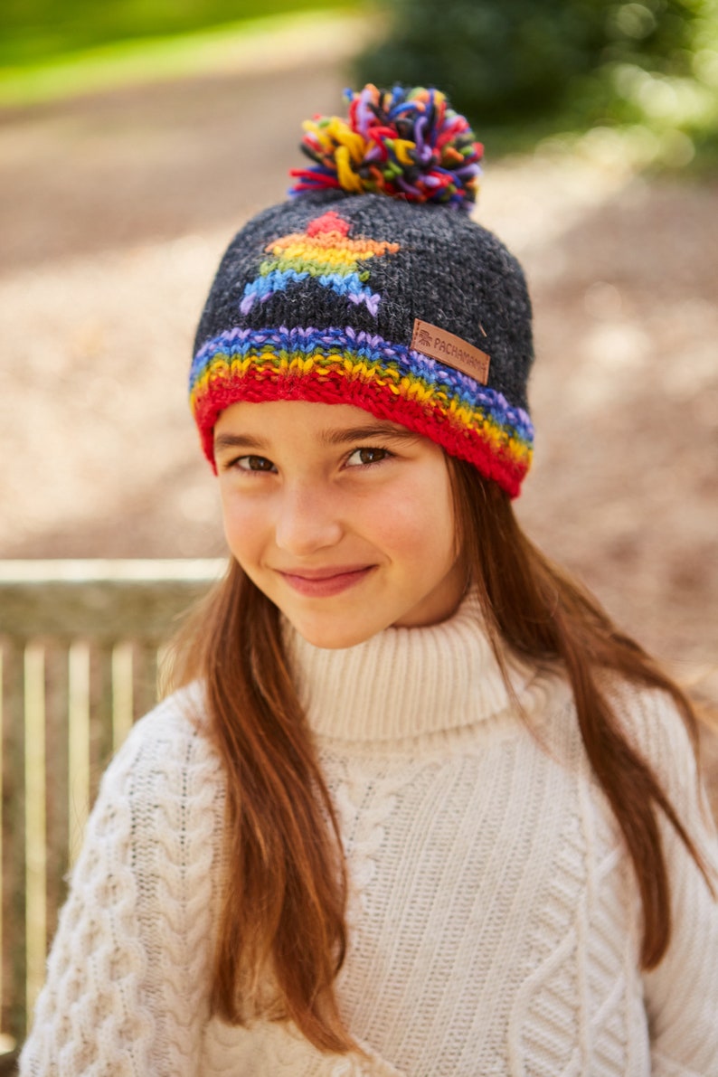 May include: A child wearing a gray knitted hat with a rainbow star design and a colorful pom-pom on top. The hat has a red, orange, yellow, green, blue, indigo, and violet stripe around the brim. The hat has a brown leather label with the text "Pachamama" on it.
