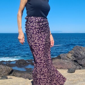 May include: A woman wearing a black tank top and a long purple skirt with a white swirl pattern. The skirt is tied at the waist and flows down to her ankles. She is standing on a rocky beach with the ocean in the background.