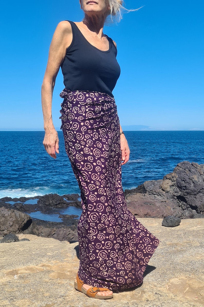 May include: A woman wearing a black tank top and a long purple skirt with a white swirl pattern. The skirt is tied at the waist and flows down to her ankles. She is standing on a rocky beach with the ocean in the background.