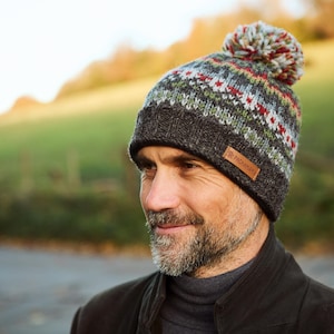 May include: A gray and green patterned knit beanie with a brown pom pom on top. The beanie has a brown leather label with the text "The Wool Company" on it.