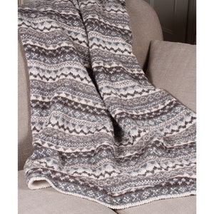 K&ouml;nnte beinhalten: Eine gestrickte Decke mit geometrischem Muster in Grau-, Braun- und Cremet&ouml;nen. Die Decke ist &uuml;ber einem neutralfarbenen Sessel drapiert und zeigt ihr strukturiertes Design. Die R&auml;nder der Decke sind mit einem wei&szlig;en Rand versehen.