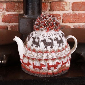 Könnte beinhalten: Eine weiße Teekanne mit einer gestrickten Teekanne in einem rot-weiß-braunen Muster. Die Teekanne hat einen Bommel oben und zeigt ein sich wiederholendes Muster aus kleinen Tieren.