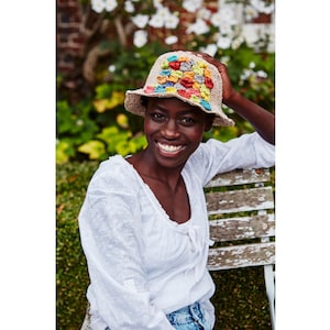 May include: A woman wearing a white crochet top and blue jeans smiles while wearing a crocheted hat with a colorful patchwork design.