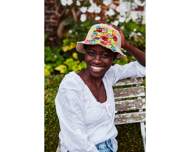 May include: A woman wearing a white crochet top and blue jeans smiles while wearing a crocheted hat with a colorful patchwork design.
