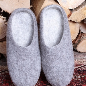May include: A pair of grey felted wool slippers. The slippers have a rounded toe and a low-cut design, with a light grey interior. They are displayed on a patterned rug with a wood background.