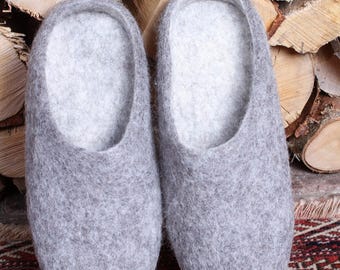 May include: A pair of grey felted wool slippers. The slippers have a rounded toe and a low-cut design, with a light grey interior. They are displayed on a patterned rug with a wood background.