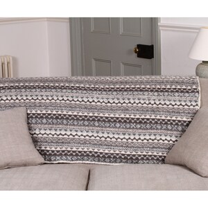 K&ouml;nnte beinhalten: Eine gestrickte Decke mit geometrischem Muster in Grau-, Wei&szlig;- und Braunt&ouml;nen, die &uuml;ber einem Sofa drapiert ist. Die Decke wird mit zwei passenden grauen Kissen pr&auml;sentiert. Das Sofa befindet sich in einem Wohnzimmer.