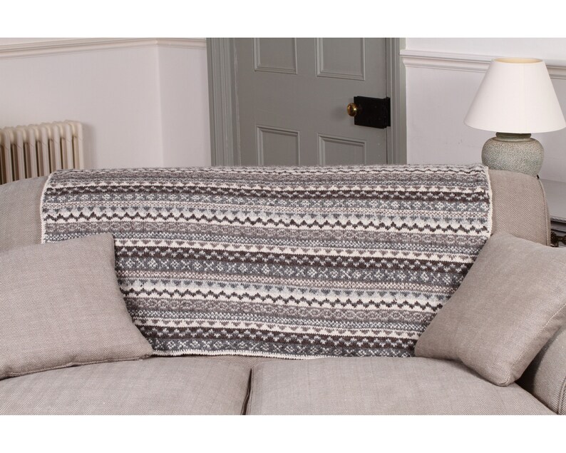 K&ouml;nnte beinhalten: Eine gestrickte Decke mit geometrischem Muster in Grau-, Wei&szlig;- und Braunt&ouml;nen, die &uuml;ber einem Sofa drapiert ist. Die Decke wird mit zwei passenden grauen Kissen pr&auml;sentiert. Das Sofa befindet sich in einem Wohnzimmer.