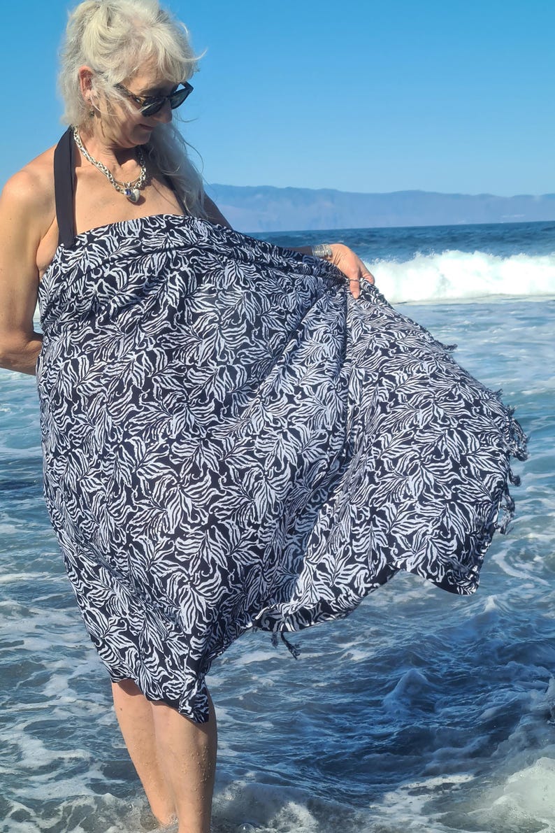 May include: A woman wearing a black and white patterned sarong on a beach. The sarong has a fringed edge and is made of a lightweight fabric.