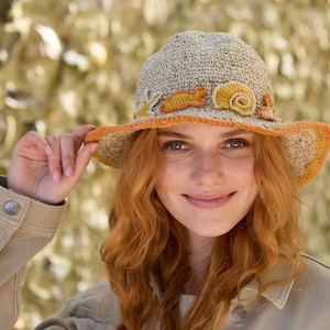 May include: A woman wearing a beige crocheted sun hat with orange and yellow accents. The hat has a wide brim and a decorative flower design.