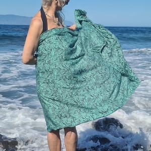 May include: A woman wearing a green and black patterned sarong on a beach. The sarong is wrapped around her waist and tied at her side. She is standing on a rocky beach with waves crashing in the background.