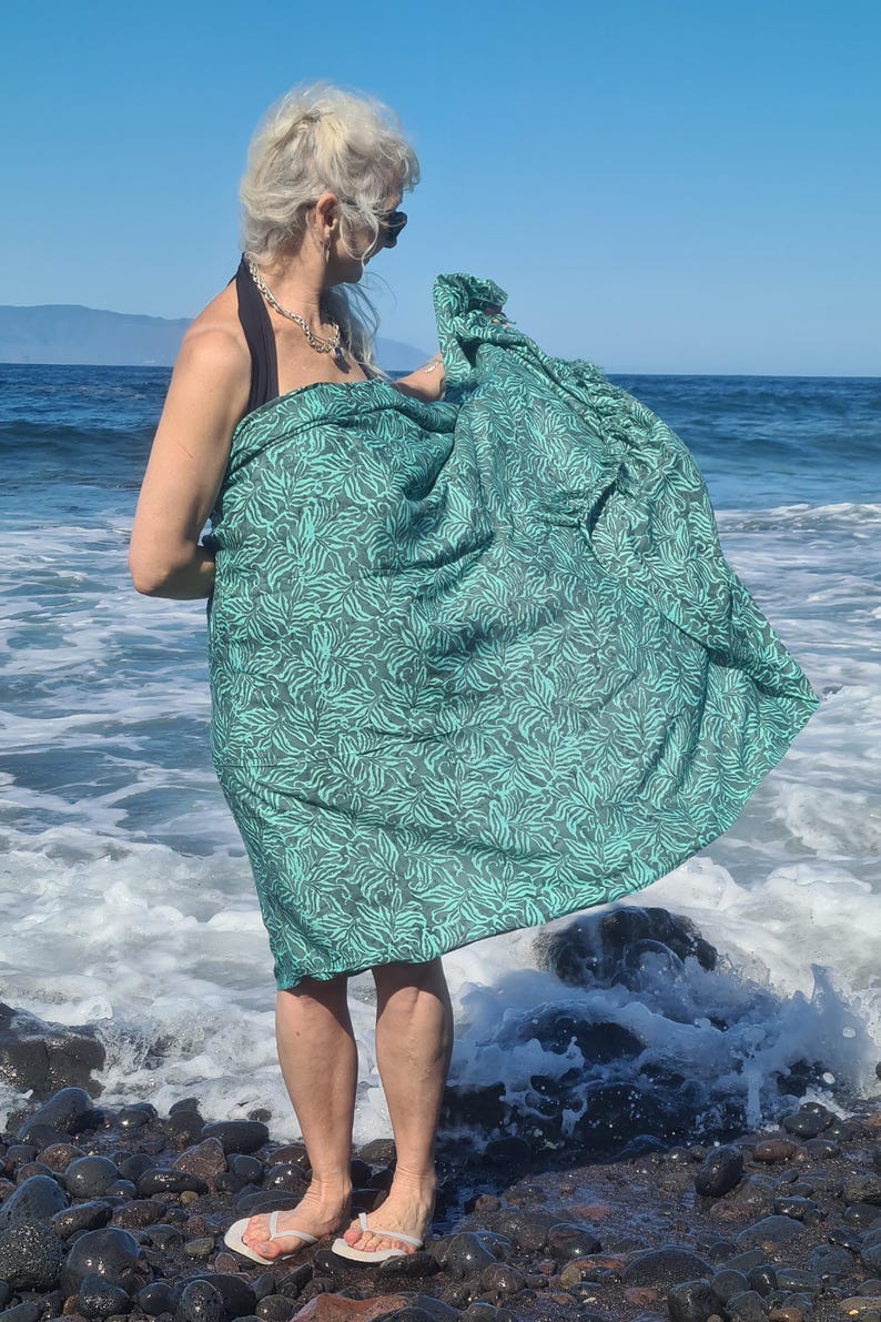 May include: A woman wearing a green and black patterned sarong on a beach. The sarong is wrapped around her waist and tied at her side. She is standing on a rocky beach with waves crashing in the background.