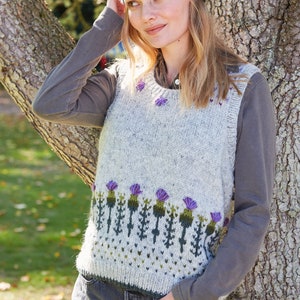 May include: A gray knitted vest with a purple and green thistle pattern. The vest has a scoop neck and sleeveless design. The model is wearing a purple and gray headband with a thistle pattern.