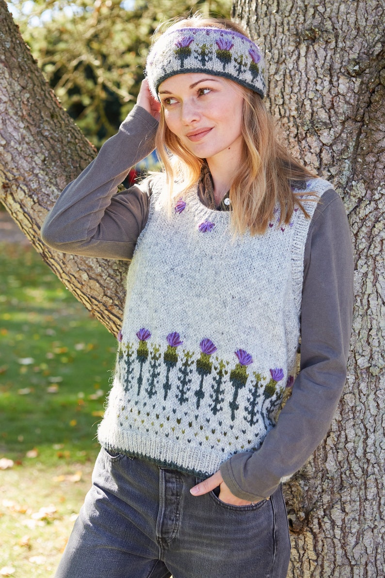 May include: A gray knitted vest with a purple and green thistle pattern. The vest has a scoop neck and sleeveless design. The model is wearing a purple and gray headband with a thistle pattern.