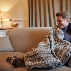 K&ouml;nnte beinhalten: Gem&uuml;tliche Wohnzimmerszene mit einer Person auf einem beigen Sofa, bedeckt mit einer gemusterten Decke. Die Decke hat ein wei&szlig;es und graues Design. Ein passendes Kissen und gemusterte Socken sind ebenfalls zu sehen.