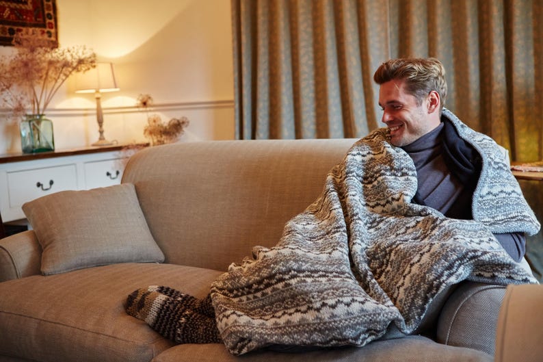 K&ouml;nnte beinhalten: Gem&uuml;tliche Wohnzimmerszene mit einer Person auf einem beigen Sofa, bedeckt mit einer gemusterten Decke. Die Decke hat ein wei&szlig;es und graues Design. Ein passendes Kissen und gemusterte Socken sind ebenfalls zu sehen.