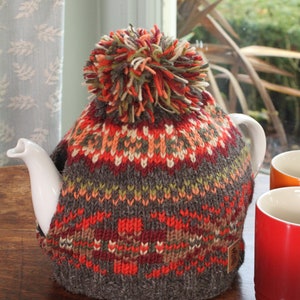 Può includere: Un copriteiera in maglia con un motivo geometrico colorato e un pompon soffice in cima. Il copriteiera è marrone, rosso, arancione e verde.