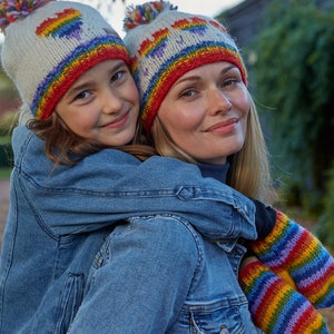 May include: A woman and a child wearing matching rainbow knit hats and mittens. The woman is wearing a denim jacket and the child is wearing a denim jacket.