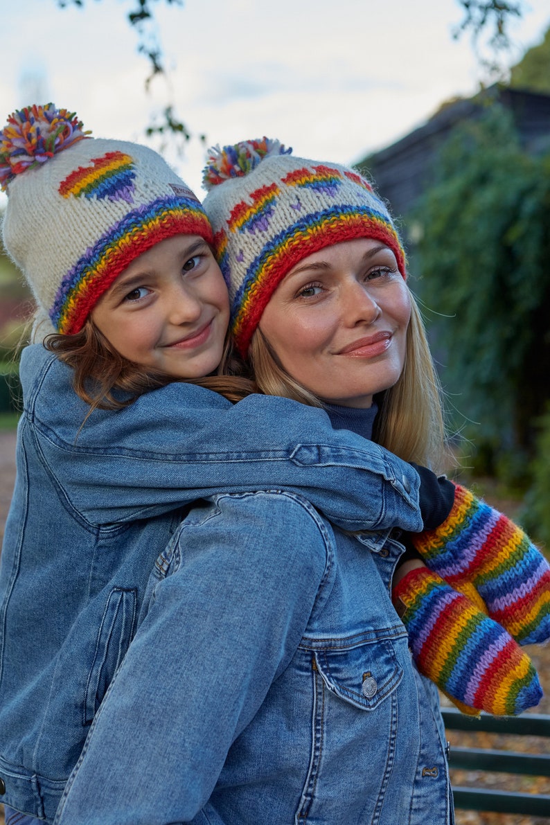 May include: A woman and a child wearing matching rainbow knit hats and mittens. The woman is wearing a denim jacket and the child is wearing a denim jacket.