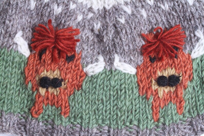 Puede incluir: Un gorro de punto con un patr&oacute;n gris, blanco y verde. Dos alces rojos y naranjas est&aacute;n bordados en el gorro.