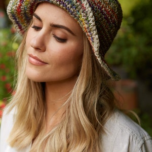 May include: A woman wearing a crocheted bucket hat with a multi-colored stripe pattern. The hat is made of natural fibers and has a relaxed, bohemian style.
