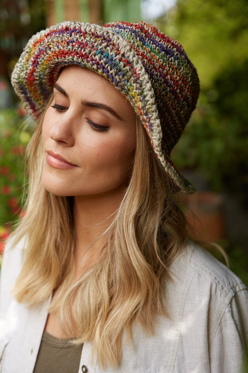 May include: A woman wearing a crocheted bucket hat with a multi-colored stripe pattern. The hat is made of natural fibers and has a relaxed, bohemian style.