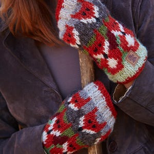 May include: A pair of red, gray, green, and white knit mittens with a fox pattern. The mittens are made of wool and have a small brown leather tag with a symbol on it.