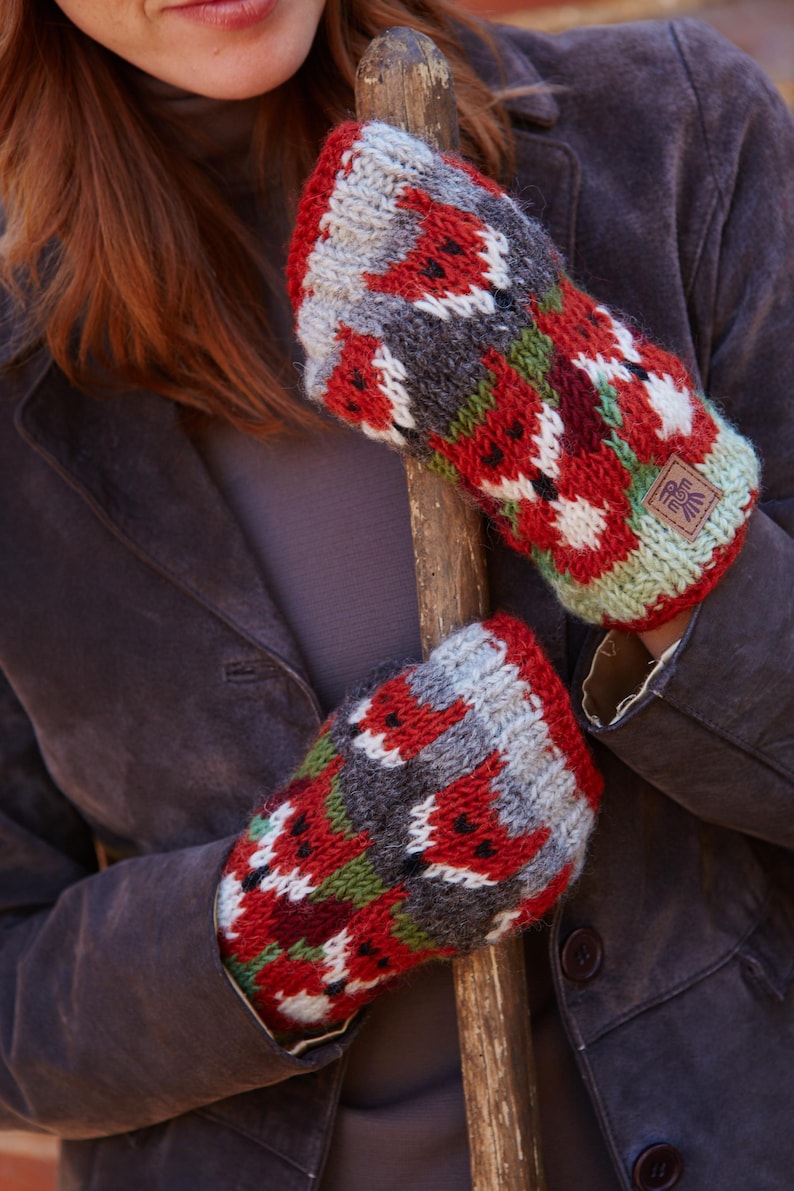 May include: A pair of red, gray, green, and white knit mittens with a fox pattern. The mittens are made of wool and have a small brown leather tag with a symbol on it.