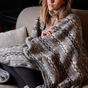 K&ouml;nnte beinhalten: Eine Person sitzt auf einem Sofa und ist in eine Strickdecke geh&uuml;llt. Die Decke hat ein Muster aus grauen, wei&szlig;en und braunen Zickzacklinien. Sie tr&auml;gt einen Rollkragenpullover und dunkle Leggings. Die Szene vermittelt Komfort und W&auml;rme.