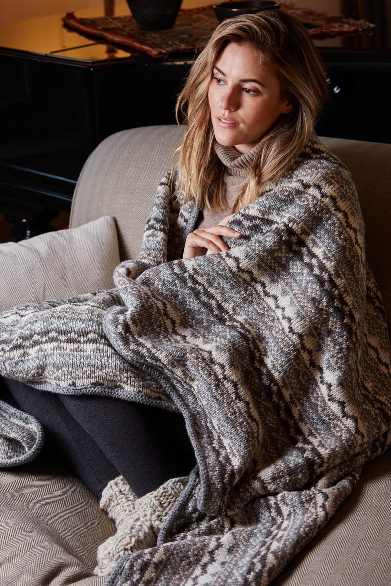 K&ouml;nnte beinhalten: Eine Person sitzt auf einem Sofa und ist in eine Strickdecke geh&uuml;llt. Die Decke hat ein Muster aus grauen, wei&szlig;en und braunen Zickzacklinien. Sie tr&auml;gt einen Rollkragenpullover und dunkle Leggings. Die Szene vermittelt Komfort und W&auml;rme.