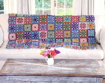 Könnte beinhalten: Eine bunte gehäkelte Wolldecke mit Blumenmuster, bestehend aus Quadraten in verschiedenen Farben, darunter Rosa, Blau, Grün, Gelb und Orange. Die Decke liegt auf einem weißen Sofa mit einem Holztisch davor.