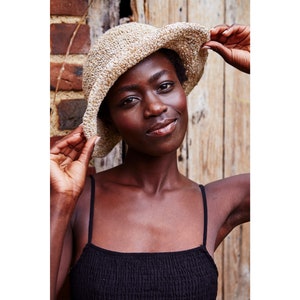 May include: A woman wearing a beige crocheted bucket hat and a black sleeveless top. She is standing in front of a wooden wall.