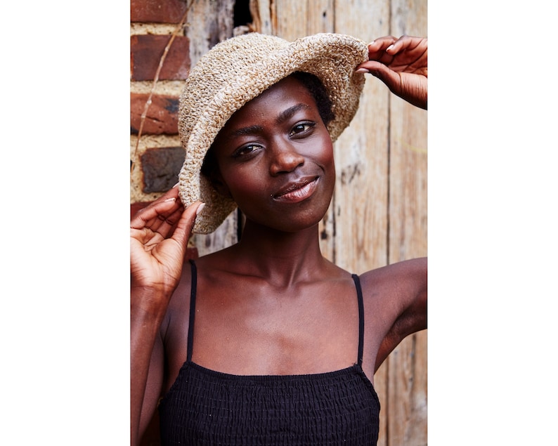 May include: A woman wearing a beige crocheted bucket hat and a black sleeveless top. She is standing in front of a wooden wall.