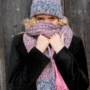 Puede incluir: Una mujer con un abrigo negro, un gorro de punto a rayas rosa y gris con un pompón rosa y una bufanda de punto a rayas rosa y gris. La bufanda está envuelta alrededor de su cuello y ella está sosteniendo los extremos de la bufanda en sus manos.
