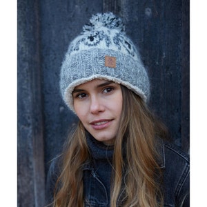 May include: A grey and white knitted beanie with a pom pom on top. The beanie has a white fleece lining and a small brown leather patch with the word "Alpaca" on it.
