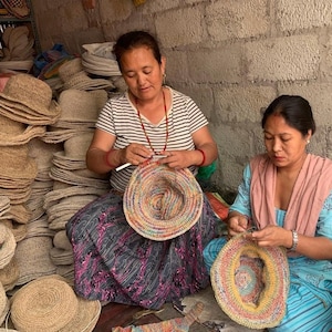 May include: Two people are sitting on the floor and making colorful crocheted hats. The hats are made with a variety of colors, including red, orange, yellow, green, blue, and purple. The hats are being made with a crochet hook and yarn. There are many other hats stacked in the background.