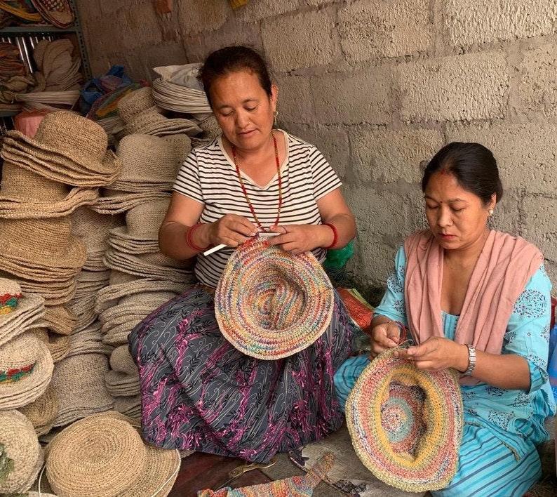 May include: Two people are sitting on the floor and making colorful crocheted hats. The hats are made with a variety of colors, including red, orange, yellow, green, blue, and purple. The hats are being made with a crochet hook and yarn. There are many other hats stacked in the background.