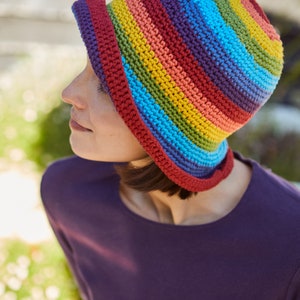 May include: A woman wearing a crocheted rainbow striped hat. The hat has a wide brim and is made with a variety of colorful yarns.
