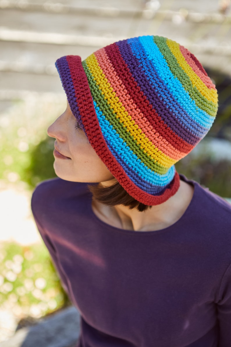 May include: A woman wearing a crocheted rainbow striped hat. The hat has a wide brim and is made with a variety of colorful yarns.