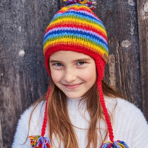May include: A young girl wearing a rainbow striped knit hat with a red pom pom on top and matching red ear flaps. The hat has a brown leather tag on the side.