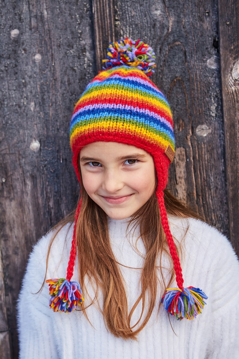 May include: A young girl wearing a rainbow striped knit hat with a red pom pom on top and matching red ear flaps. The hat has a brown leather tag on the side.