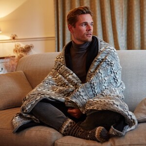 K&ouml;nnte beinhalten: Eine Person, eingeh&uuml;llt in eine gemusterte Decke, sitzt auf einem hellbraunen Sofa. Die Decke hat ein grau-wei&szlig;es Design. Die Person tr&auml;gt einen grauen Rollkragenpullover und eine dunkle Hose. Der Raum hat eine warme, einladende Atmosph&auml;re.