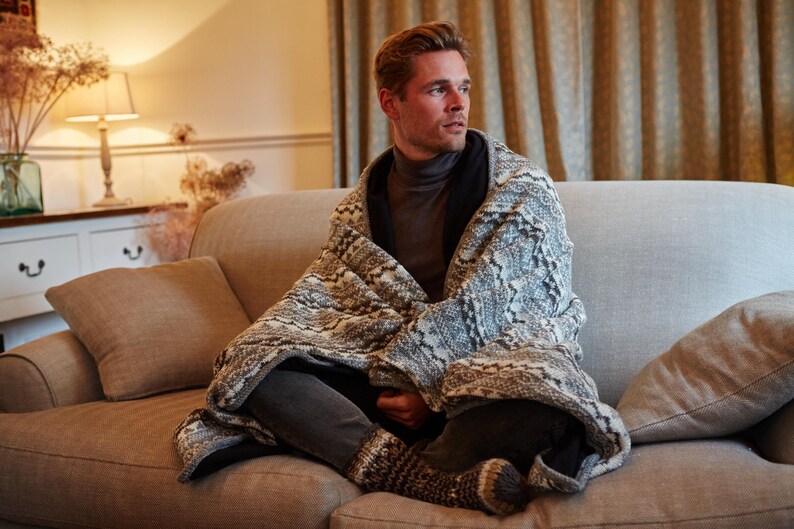 K&ouml;nnte beinhalten: Eine Person, eingeh&uuml;llt in eine gemusterte Decke, sitzt auf einem hellbraunen Sofa. Die Decke hat ein grau-wei&szlig;es Design. Die Person tr&auml;gt einen grauen Rollkragenpullover und eine dunkle Hose. Der Raum hat eine warme, einladende Atmosph&auml;re.
