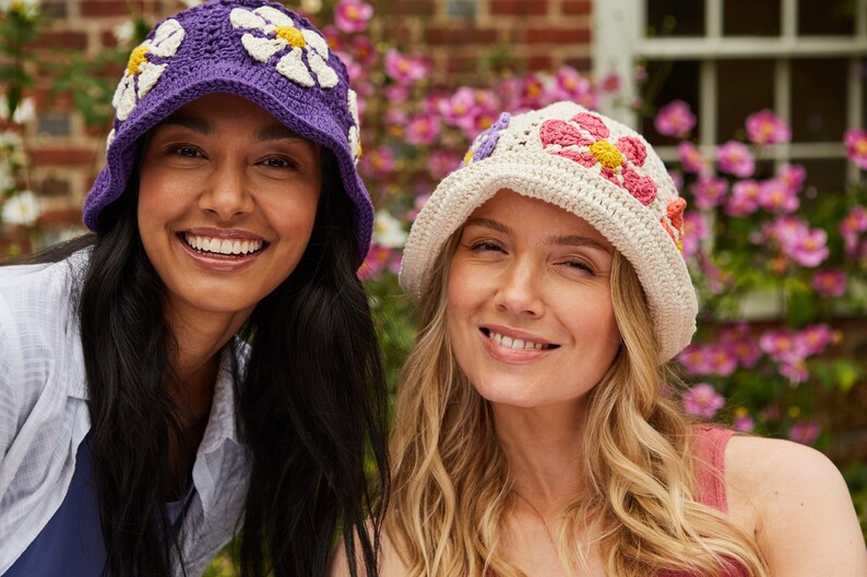 May include: Two women wearing crocheted flower hats. The woman on the left is wearing a purple hat with white flowers. The woman on the right is wearing a white hat with pink, orange, and yellow flowers.