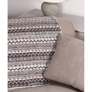 K&ouml;nnte beinhalten: Eine Strickdecke mit geometrischem Muster in Grau-, Braun- und Cremet&ouml;nen liegt &uuml;ber einem Sofa. Ein einfarbiges, hellbraunes Kissen liegt an der Decke. Im Hintergrund befindet sich eine Lampe mit wei&szlig;em Schirm.
